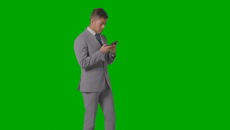 Three-Quarter-Length-Studio-Shot-Of-Businessman-In-Suit-Looking-At-Mobile-Phone-Against-Green-Screen-