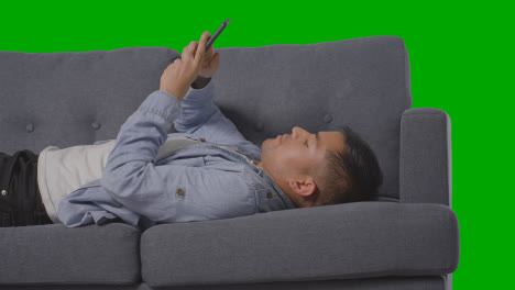 Studio-Shot-Of-Man-Lying-On-Sofa-Scrolling-Through-Mobile-Phone-Against-Green-Screen