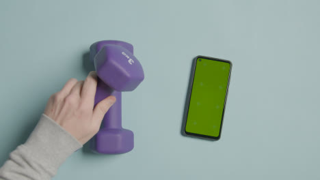 Overhead-Fitness-Studio-Shot-Of-Male-Hand-Picking-Up-Exercise-Dumbbell-Weights-Next-To-Green-Screen-Mobile-Phone-5