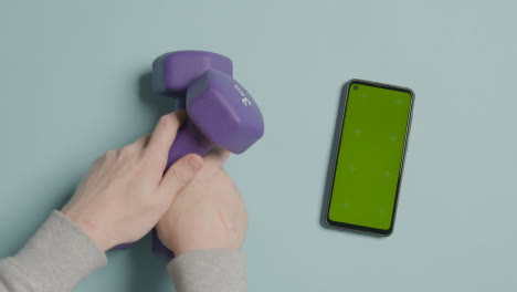 Overhead-Fitness-Studio-Shot-Of-Male-Hand-Picking-Up-Exercise-Dumbbell-Weights-Next-To-Green-Screen-Mobile-Phone-6