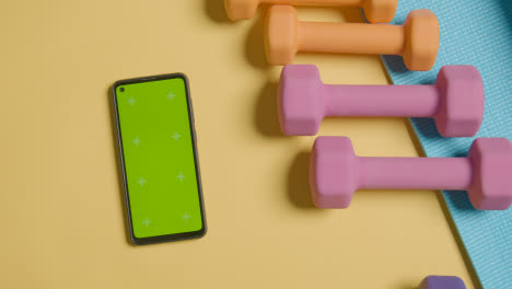Overhead-Fitness-Studio-Shot-Of-Hand-Picking-Up-Coloured-Exercise-Dumbbell-Weight-Next-To-Green-Screen-Mobile-Phone