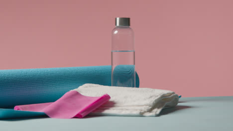 Fitness-Studio-Shot-Of-Water-Bottle-With-Exercise-Mat-And-Towel-Against-Pink-Background