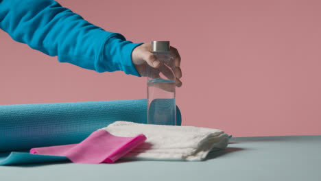 Fitness-Studio-Shot-Of-Person-Picking-Up-Water-Bottle-With-Exercise-Mat-And-Towel-Against-Pink-Background