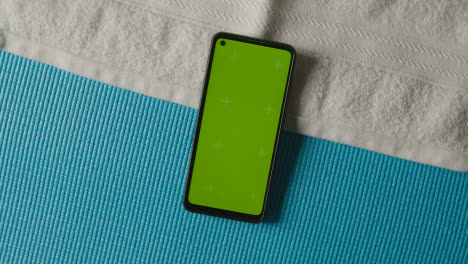 Overhead-Studio-Fitness-Shot-Of-Rotating-Green-Screen-Mobile-Phone-With-Trainers-And-Towel-On-Blue-Exercise-Mat