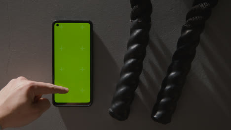 Overhead-Studio-Fitness-Shot-Of-Person-Using-Green-Screen-Mobile-Phone-Next-To-Gym-Battle-Ropes-On-Grey-Background
