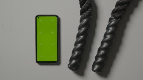 Overhead-Studio-Fitness-Shot-Of-Person-Using-Green-Screen-Mobile-Phone-Next-To-Gym-Battle-Ropes-On-Grey-Background-1