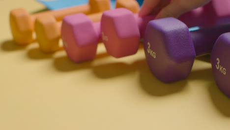 Studio-Fitness-Shot-With-Hand-Putting-Down-Colourful-Gym-Weights-In-A-Line-On-Yellow-Background