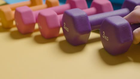 Foto-De-Estudio-De-Fitness-Con-La-Mano-Recogiendo-Pesas-De-Gimnasio-Coloridas-En-Una-Línea-Sobre-Fondo-Amarillo