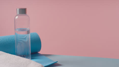 Fitness-Studio-Shot-Of-Person-Picking-Up-Water-Bottle-With-Exercise-Mat-And-Towel-Against-Pink-Background-1