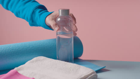 Fitness-Studio-Shot-Of-Person-Picking-Up-Water-Bottle-With-Exercise-Mat-And-Towel-Against-Pink-Background-2
