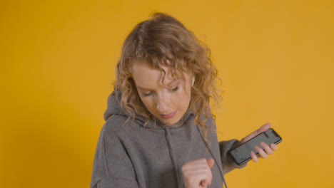 Studio-Shot-Of-Young-Woman-Listening-To-Music-On-Mobile-Phone-And-Dancing-Against-Yellow-Background-1