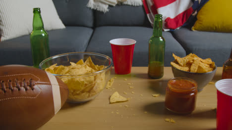 Lounge-Zu-Hause-Einer-Person,-Die-Ein-American-Football-Spiel-Im-Fernsehen-Mit-Getränken,-Snacks-Und-Ball-Sieht