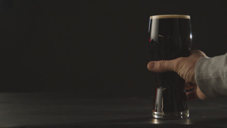 Person-Picking-Up-Pint-Of-Irish-Stout-In-Glass-Against-Black-Studio-Background-To-Celebrate-St-Patricks-Day-2