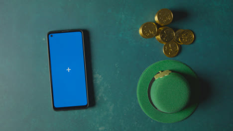 Overhead-Studio-Shot-Of-Green-Leprechaun-Top-Hat-With-Gold-Coins-And-Blue-Screen-Mobile-Phone-To-Celebrate-St-Patricks-Day