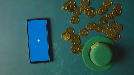Overhead-Studio-Shot-Of-Green-Leprechaun-Top-Hat-With-Gold-Coins-And-Blue-Screen-Mobile-Phone-To-Celebrate-St-Patricks-Day-3