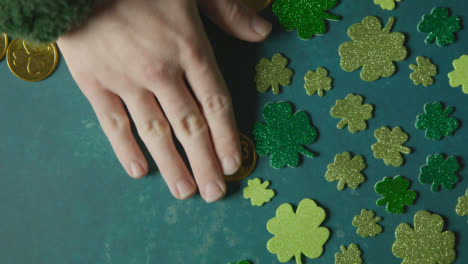 Overhead-Studio-Shot-Of-Hand-Collecting-Gold-Coins-Next-To-Shamrock-Shapes-On-Background-To-Celebrate-St-Patricks-Day