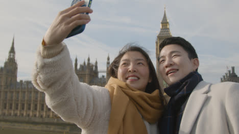 Joven-Pareja-Asiática-De-Vacaciones-Posando-Para-Selfie-Frente-A-Las-Casas-Del-Parlamento-En-Londres,-Reino-Unido
