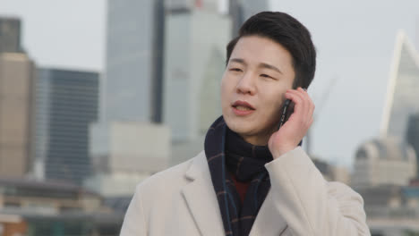 Young-Asian-Businessman-Answering-Call-On-Mobile-Phone-With-London-City-Skyline-In-Background