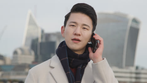 Young-Asian-Businessman-Talking-On-Mobile-Phone-With-London-City-Skyline-In-Background