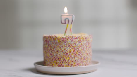 Studio-Shot-Birthday-Cake-Covered-With-Decorations-And-Lit-Candle-Celebrating-Seventh-Birthday-Being-Blown-Out