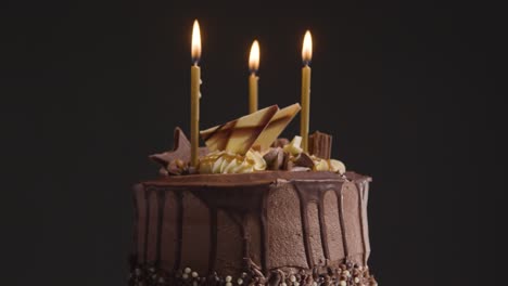 Nahaufnahme-Studioaufnahme-Von-Dekoriertem-Schokoladengeburtstagskuchen-Mit-Kerzen,-Die-Sich-Vor-Schwarzem-Hintergrund-Drehen