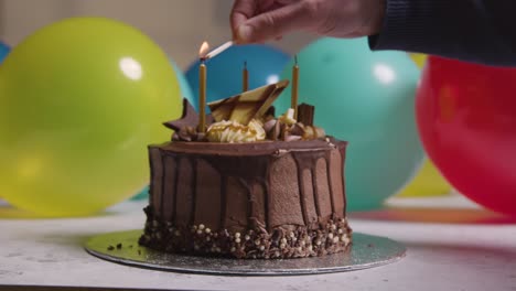 Dekorierter-Schokoladen-Geburtstagskuchen-Mit-Kerzen,-Die-Auf-Einer-Party-Mit-Luftballons-Im-Hintergrund-Angezündet-Werden