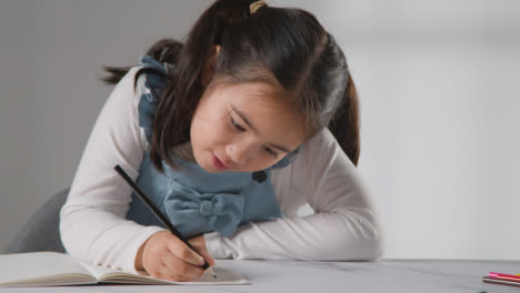 Studio-Shot-Of-Young-Girl-At-Table-Writing-In-School-Book-1