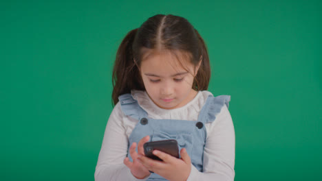 Studioporträt-Eines-Jungen-Mädchens-Im-ASD-Spektrum,-Das-Mit-Einem-Mobiltelefon-Auf-Grünem-Hintergrund-Spielt