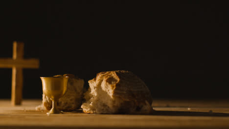 Religious-Concept-Shot-With-Chalice-Broken-Bread-Cross-And-Wine-On-Wooden-Table-Against-Black-Background-1
