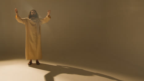 Full-Length-Studio-Portrait-Of-Man-Wearing-Robes-And-Sandals-With-Long-Hair-And-Beard-Representing-Figure-Of-Jesus-Christ-With-Arms-Outstretched