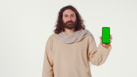 Porträt-Eines-Mannes-In-Roben-Mit-Langen-Haaren-Und-Bart,-Der-Die-Figur-Von-Jesus-Christus-Darstellt-Und-Ein-Mobiltelefon-Mit-Grünem-Bildschirm-Hält