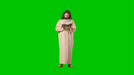 Studio-Shot-Of-Man-Wearing-Robes-And-Sandals-With-Long-Hair-And-Beard-Representing-Figure-Of-Jesus-Christ-Reading-Bible-On-Green-Screen-