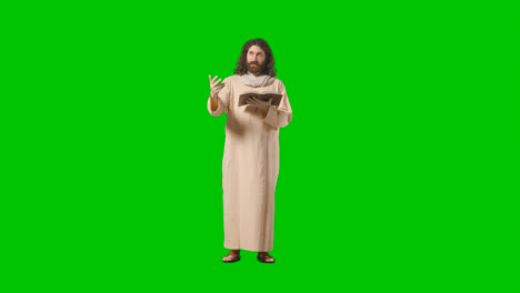 Studio-Shot-Of-Man-Wearing-Robes-And-Sandals-With-Long-Hair-And-Beard-Representing-Figure-Of-Jesus-Christ-Preaching-From-Bible-On-Green-Screen-
