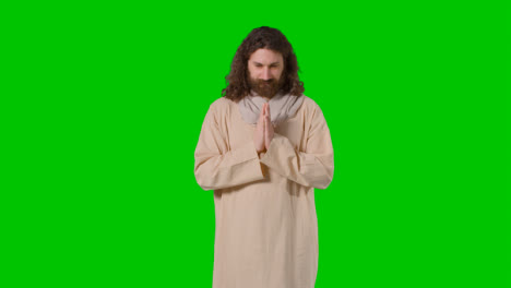 Studioaufnahme-Eines-Mannes-In-Roben-Mit-Langen-Haaren-Und-Bart,-Der-Die-Figur-Des-Betenden-Jesus-Christus-Auf-Einem-Grünen-Bildschirm-Darstellt