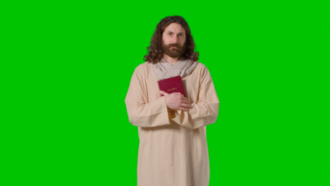 Studioaufnahme-Eines-Mannes-In-Roben-Mit-Langen-Haaren-Und-Bart,-Der-Die-Figur-Von-Jesus-Christus-Darstellt,-Der-Die-Bibel-Hält,-Auf-Grünem-Bildschirm