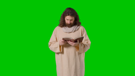Studio-Shot-Of-Man-Wearing-Robes-With-Long-Hair-And-Beard-Representing-Figure-Of-Jesus-Christ-Reading-Bible-On-Green-Screen-