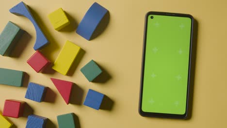Overhead-Shot-Of-Colourful-Wooden-Building-Blocks-On-Yellow-Studio-Background-With-Green-Screen-Mobile-Phone