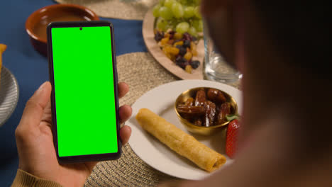Man-Looking-At-Green-Screen-Mobile-Phone-Sitting-Around-Muslim-Family-Table-At-Home-Eating-Iftar-Meal-Breaking-Daily-Fast-During-Ramadan