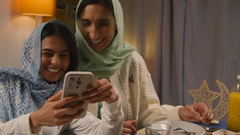 Mother-And-Daughter-Laughing-At-Mobile-Phone-Sitting-Around-Muslim-Family-Table-At-Home-Eating-Iftar-Meal-Breaking-Daily-Fast-During-Ramadan-1
