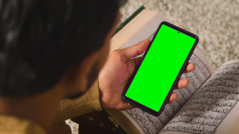 Close-Up-Of-Muslim-Man-At-Home-Sitting-On-Floor-And-Reciting-From-The-Quran-Holding-Green-Screen-Mobile-Phone