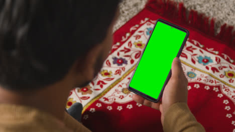 Close-Up-Of-Muslim-Man-At-Home-Sitting-On-Floor-On-Prayer-Mat-Holding-Green-Screen-Mobile-Phone