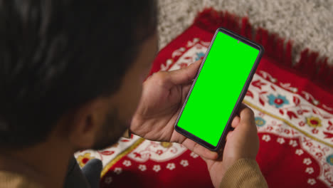 Close-Up-Of-Muslim-Man-At-Home-Sitting-On-Floor-On-Prayer-Mat-Holding-Green-Screen-Mobile-Phone-1