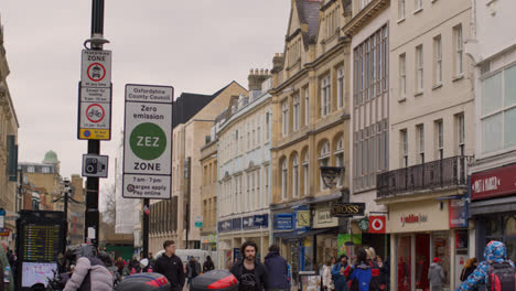 Vista-A-Lo-Largo-De-Cornmarket-Street-En-El-Centro-De-La-Ciudad-De-Oxford-Con-Tiendas-Y-Peatones-Y-Señales-De-Zona-De-Tráfico-De-Cero-Emisiones