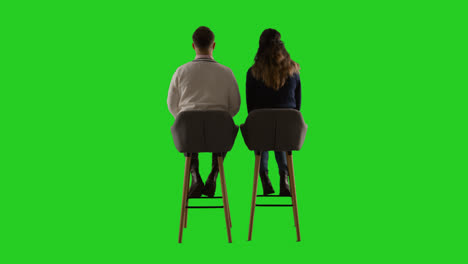 Rear-View-Studio-Shot-Of-Man-And-Woman-Sitting-On-Stools-Facing-Away-From-Camera-Against-Green-Screen