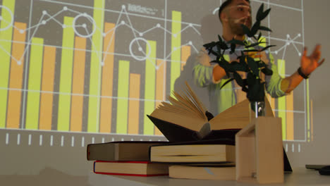 Close-Up-Of-Textbooks-On-Desk-As-Teacher-Gives-Maths-Or-Science-Lesson-In-School-Standing-At-The-Front-Of-Class-With-Projection-Of-Graph