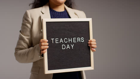 Close-Up-Studio-Shot-Of-Female-Teacher-Standing-Against-Grey-Background-Holding-Notice-Board-Reading-Teachers-Day-3