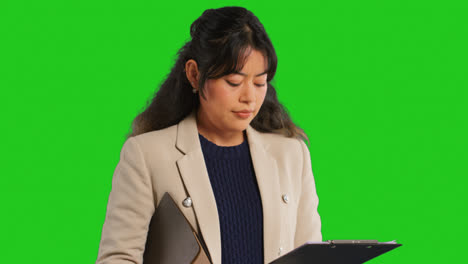 Close-Up-Studio-Portrait-Of-Female-Teacher-Or-Businesswoman-Standing-Against-Green-Screen-Looking-At-Clipboard