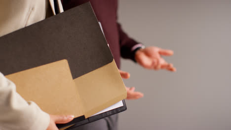 Close-Up-Studio-Shot-Of-Male-And-Female-Teachers-Against-Grey-Background-Holding-Folder-Under-Arm-Walking-Together