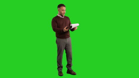 Full-Length-Studio-Portrait-Of-Mature-Male-Teacher-Or-Businessman-Standing-Against-Green-Screen-Looking-At-Clipboard