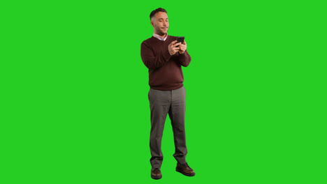 Full-Length-Studio-Portrait-Of-Mature-Male-Teacher-Or-Businessman-Standing-Against-Green-Screen-Using-Mobile-Phone-To-Message-Or-Browse-Online